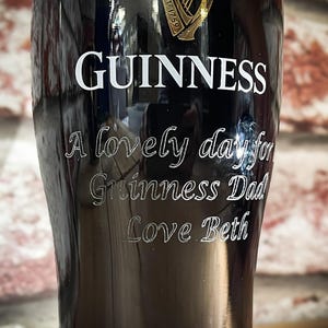 May include: A black Guinness pint glass with white lettering. The word "GUINNESS" is at the top, with the phrases "A lovely day for Guinness Dad Love Beth" etched below. The glass is set against a brick wall.