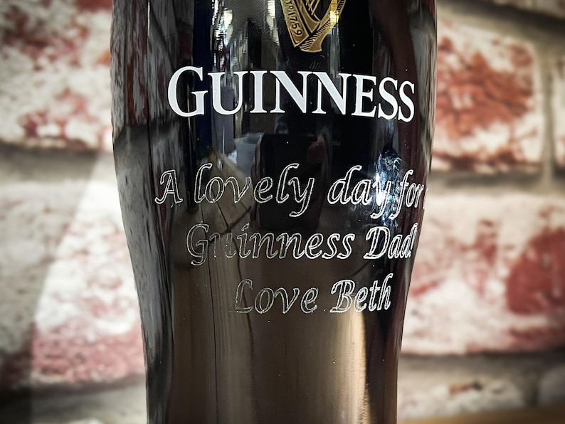 May include: A black Guinness pint glass with white lettering. The word "GUINNESS" is at the top, with the phrases "A lovely day for Guinness Dad Love Beth" etched below. The glass is set against a brick wall.