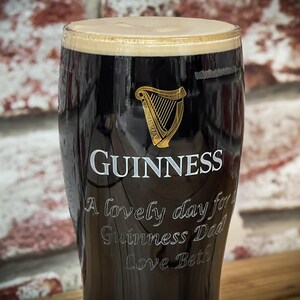 May include: A pint glass filled with dark stout, topped with a creamy head. The Guinness logo and a golden harp are visible. Text on the glass reads "A lovely day for Guinness Dad Love Beth."