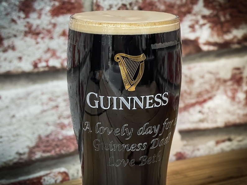 May include: A pint glass filled with dark stout, topped with a creamy head. The Guinness logo and a golden harp are visible. Text on the glass reads "A lovely day for Guinness Dad Love Beth."