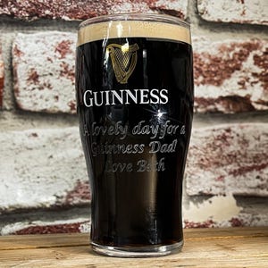 Puede incluir: Un vaso de pinta lleno de cerveza negra oscura, coronado con espuma cremosa. El logotipo de Guinness y la inscripción "A lovely day for a Guinness Dad Love Beth" están grabados. El vaso está sobre una superficie de madera.