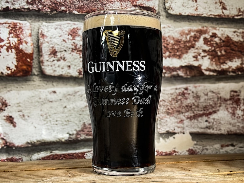 May include: A pint glass filled with dark stout, topped with a creamy head. The Guinness logo and the words "A lovely day for a Guinness Dad Love Beth" are etched on the glass. The glass sits on a wooden surface.