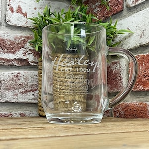 Könnte beinhalten: Eine klare Glas-Tasse mit Henkel. Die Tasse ist mit dem Text "Healey Est. 1980" graviert.