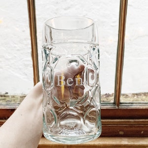 Könnte beinhalten: Ein klarer Bierkrug aus Glas mit einem strukturierten, blasenartigen Muster. Der Name "Ben" ist auf der Vorderseite eingraviert. Der Krug wird vor einem Fenster mit Holzrahmen gehalten.