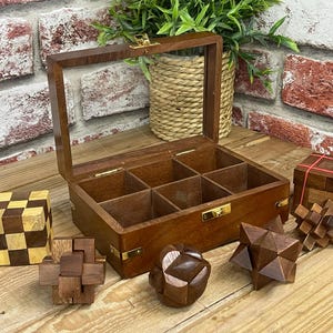 Könnte beinhalten: Eine Holzteebox mit Glasdeckel, geöffnet, um mehrere Fächer freizugeben. Mehrere Holzpuzzles in verschiedenen Formen und Farben sind um die Box herum angeordnet, darunter ein Würfel und sternförmige Puzzles. Eine dekorative Pflanze befindet sich im Hintergrund.