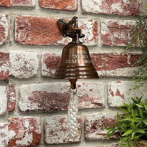 May include: A copper bell with a rope pull hanging on a brick wall. The bell has the text "Ring to buy staff drinks" engraved on it.