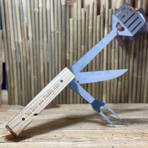 May include: A grilling tool set with a wooden handle, featuring a spatula, knife, and fork. The handle is engraved with the text "We love you Daddy xxx".