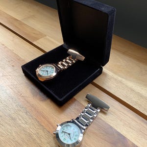 May include: Two nurse fob watches, one in a black velvet box. The watches have a round face with a white background and green numerals. One watch is gold-toned, the other is silver-toned. The watches are on a wooden surface.