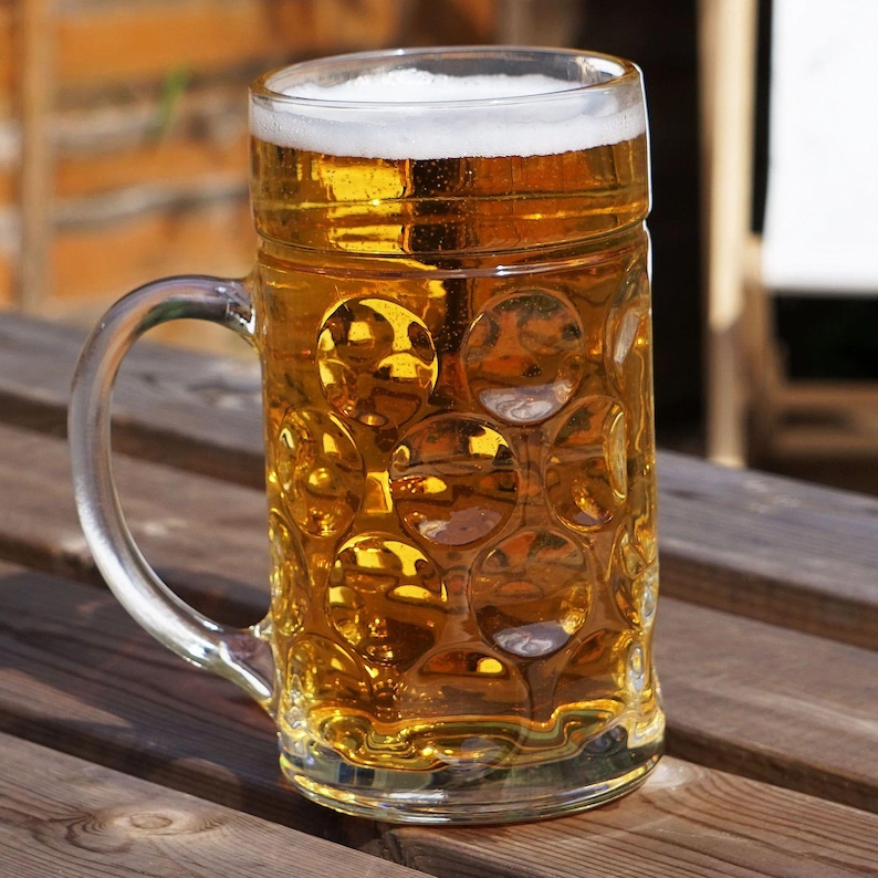 May include: A large glass beer mug filled with golden beer and a foamy white head. The clear glass mug has a handle and a textured pattern. The mug sits on a wooden table, with a blurred background.