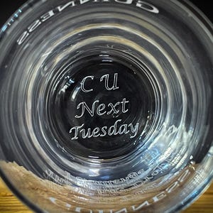 May include: Clear glass with the words "CU Next Tuesday" etched in white script. The glass has a wide rim and a slightly textured surface. The image is taken from a low angle, looking up into the glass.