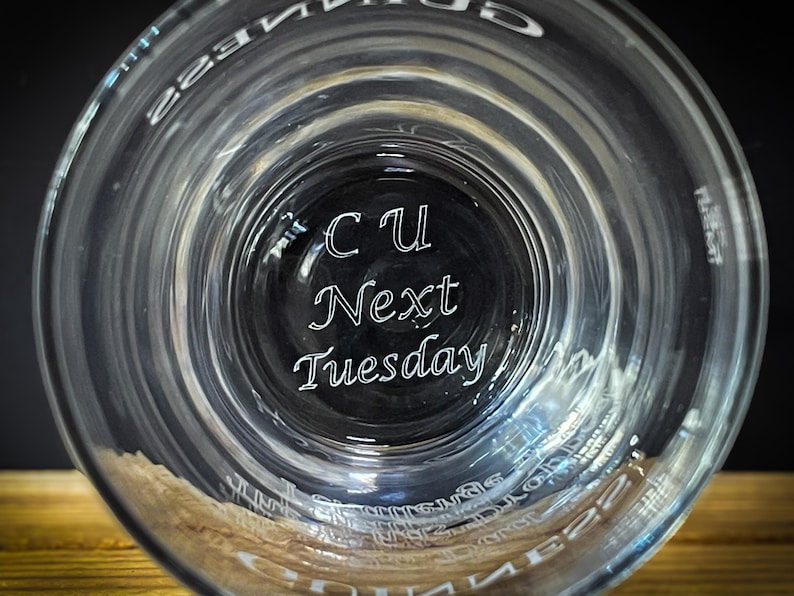 May include: Clear glass with the words "CU Next Tuesday" etched in white script. The glass has a wide rim and a slightly textured surface. The image is taken from a low angle, looking up into the glass.