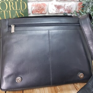 May include: Black leather messenger bag with a zippered pocket and a strap. The bag has two silver metal accents on the front.