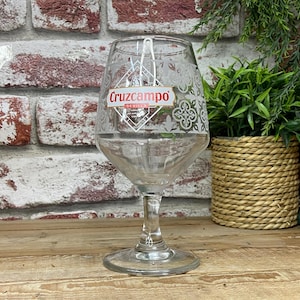 May include: A clear glass beer goblet with a stem and a wide bowl. The glass has the "Cruzcampo Sevilla" logo and decorative patterns. The glass is on a wooden surface.
