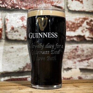 May include: A pint glass filled with dark stout, featuring the Guinness logo and the text "A lovely day for a Guinness Dad Love Beth." The glass is set against a brick wall background.
