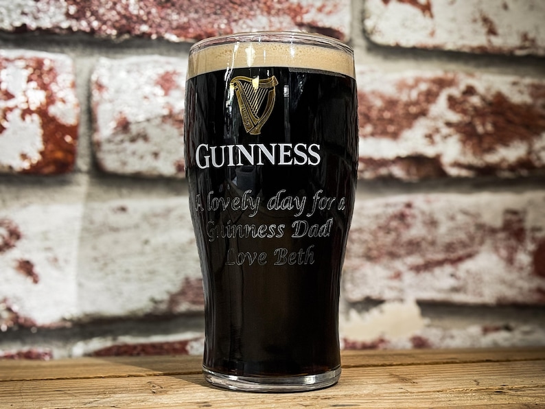 May include: A pint glass filled with dark stout, featuring the Guinness logo and the text "A lovely day for a Guinness Dad Love Beth." The glass is set against a brick wall background.