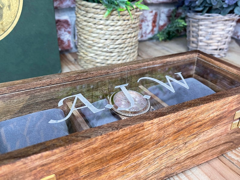 May include: A wooden watch box with a glass lid. The box has a grey velvet interior and is engraved with the initials "A.J.W.".