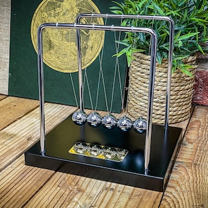 Op de afbeelding: Een Newton's cradle van chroom met vijf zilveren ballen die aan touwtjes hangen. De ballen zijn opgehangen aan een zwarte basis met een gouden accent.