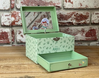 May include: A light green jewellery box with a hinged lid and a pull-out drawer. The box features a mirror inside the lid and a decorative spinning figurine. The box is decorated with leaf patterns and the name "Ariel".