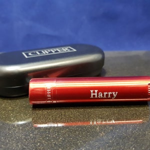 May include: Red metal lighter with the word "Clipper" engraved on the side and the name "Harry" engraved on the top. The lighter is in a black case.