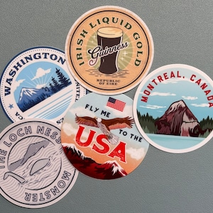 May include: A collection of round stickers featuring travel-themed designs. One sticker displays a pint of Guinness with "Irish Liquid Gold." Others showcase "Washington," "The Loch Ness Monster," "Fly Me to the USA," and "Montreal, Canada."