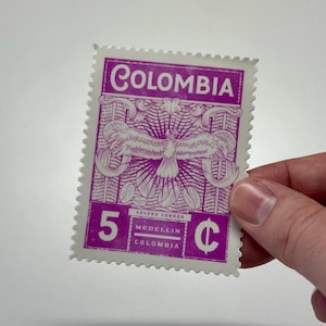 以下が含まれることがあります： コロンビアの紫と白の長方形の切手。鷲のイラスト、コーヒー豆、COLOMBIA、5、Cの文字が描かれています。切手には、小さな白い文字でVELERO CORREO、MEDELLIN、COLOMBIAという言葉が印刷されています。