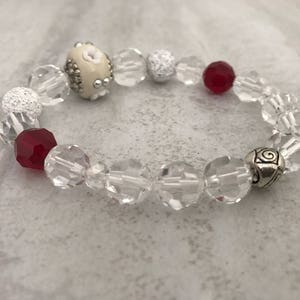 Op de afbeelding: Een kralenarmband met heldere, facetgeslepen kralen, afgewisseld met rode en zilveren accentkralen. Een crèmekleurige kraal met bloemendetails en zilveren accenten voegt een unieke touch toe. De armband is geregen op een elastiek.