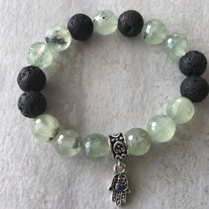 Può includere: Bracciale con perline alternate verde chiaro e nere. Le perline verde chiaro hanno un aspetto sfaccettato. Un ciondolo d'argento con un design a mano di Hamsa pende dal bracciale.