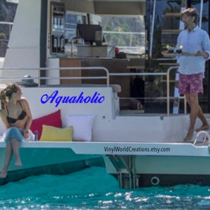 May include: A woman in a black bikini top and a blue skirt sits on a white deck of a boat. The boat is docked in the water. The word "Aquaholic" is written in blue cursive on the side of the boat. The text "VinylWorldCreations.etsy.com" is on the deck of the boat.