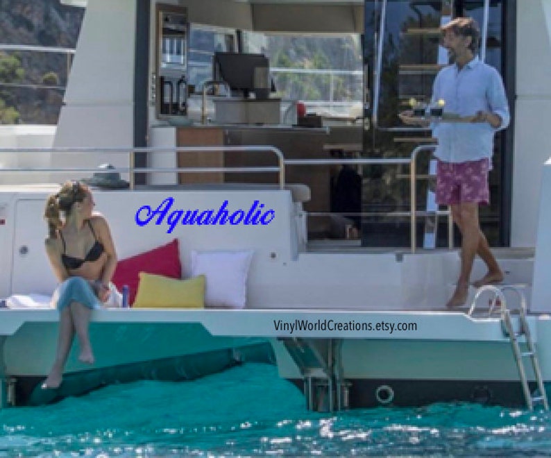 May include: A woman in a black bikini top and a blue skirt sits on a white deck of a boat. The boat is docked in the water. The word "Aquaholic" is written in blue cursive on the side of the boat. The text "VinylWorldCreations.etsy.com" is on the deck of the boat.