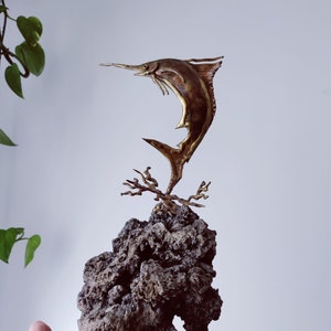 May include: A gold-colored metal sculpture of a marlin jumping out of the water, mounted on a dark gray rock base.