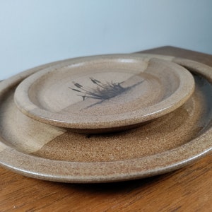 May include: Two ceramic plates, one nested inside the other. The smaller plate has a brown and white glaze with a black silhouette of a plant design. The larger plate has a brown and white glaze.