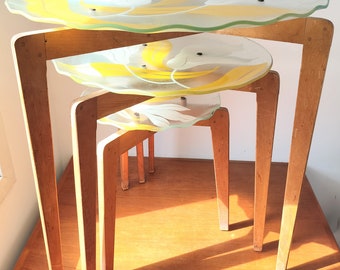 May include: A set of three nesting tables with wooden legs and glass tops. The glass tops feature a yellow and white floral design.