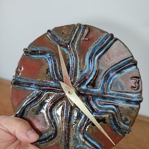 May include: A ceramic clock with a unique design featuring a swirling pattern of blue and brown glaze. The clock has a gold-colored hour hand and minute hand.