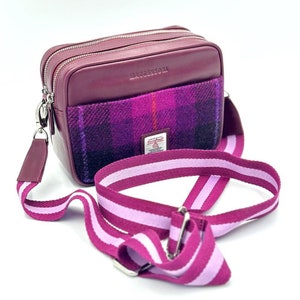 Könnte beinhalten: Eine bordeauxrote Leder-Umhängetasche mit einer karierten Stofftasche und einem verstellbaren pinkfarbenen und bordeauxroten Schultergurt. Die Tasche hat einen Reißverschluss und der Text "HARRIS TWEED" ist auf der Stofftasche sichtbar.