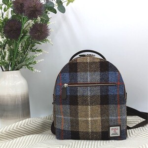 May include: A brown, blue, and orange plaid backpack with a brown leather zipper pull and a brown leather strap. The backpack has a small pocket on the front with a zipper.
