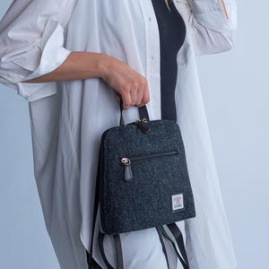 May include: A small, dark gray backpack with a textured pattern and a zippered front pocket. The bag has black straps and a small handle. The background is a plain white.