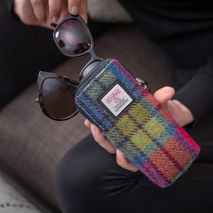 May include: A blue, green, red and yellow plaid glasses case with a zipper closure. The case has a black zipper pull and a small white label with the text "Harris Tweed" on it. The case is made of tweed fabric.