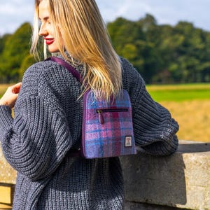 May include: A small, purple and blue plaid backpack with a zipper closure and a small square patch. The backpack has a purple strap and is worn over the shoulders. The person is wearing a gray sweater.