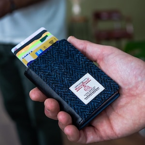 May include: A blue herringbone pattern cardholder with a black leather trim. The cardholder is filled with credit cards and features a white label with the text "Harris Tweed".