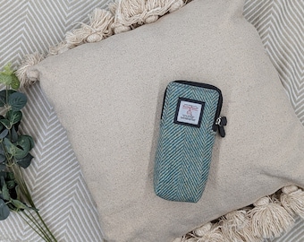 May include: A light green and white tweed phone case with a zipper closure sits on a cream-colored pillow with fringe trim. The pillow is on top of a beige and white herringbone patterned throw blanket.
