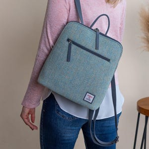 May include: A teal and grey herringbone pattern backpack with a top handle and a front zipped pocket. The backpack has grey straps and a small square label. The bag is worn over the shoulder.