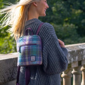 May include: A small, square backpack with a plaid pattern in shades of blue, purple, and green. The backpack has a purple zip and straps. The bag is worn over the shoulders. The background is blurred green foliage.