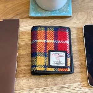 Puede incluir: Una cartera de cuadros rojos, amarillos y azules con ribete de cuero negro. La cartera tiene una etiqueta que dice "Harris Tweed" con las palabras "Outer Hebrides of Scotland" y "100% Pure New Wool" debajo.