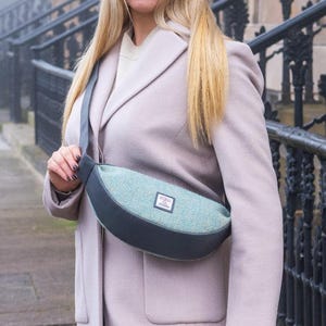 May include: A woman wearing a light pink coat and carrying a teal and gray crossbody bag. The bag has a rectangular patch on the front. The background features a wrought iron fence and a building.