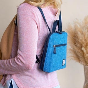May include: A small, blue backpack with a textured pattern and a zippered front pocket. The backpack has black straps and a small tag. The person is wearing a pink long-sleeve shirt, blue jeans, and a tan scarf.