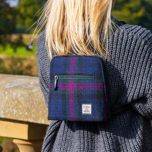 May include: A blue and purple plaid crossbody bag with a green zipper and a small logo patch. The bag is worn over a gray sweater. The background features a stone wall and trees.