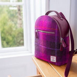 May include: A purple and pink plaid backpack with a burgundy leather trim and a matching purple and pink plaid cosmetic bag. The backpack has a zippered pocket on the front and a long strap.