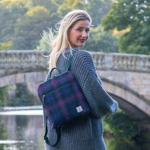 May include: A backpack in a blue, purple, and green plaid design. The person is wearing a grey jumper and blue jeans. The background shows a stone bridge over a river and green trees. The backpack has a green handle.
