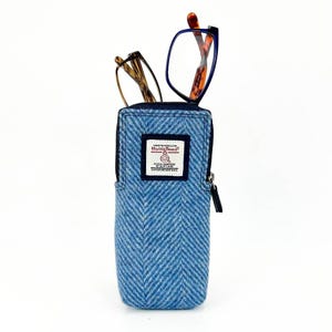 Puede incluir: Un estuche para gafas de tweed azul y blanco con un patrón de espiga y cierre de cremallera. El estuche tiene una pequeña etiqueta que dice "Harris Tweed" con un pequeño logotipo de una oveja.