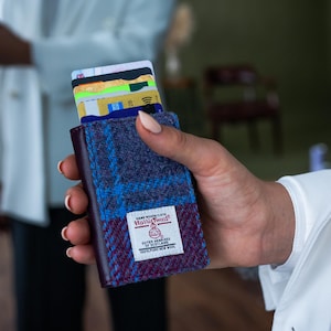 May include: A Harris Tweed wallet, showcasing a blend of purple, blue, and burgundy woven fabric. The wallet is filled with various credit cards. The Harris Tweed label is visible. The wallet is held in a person's hand.